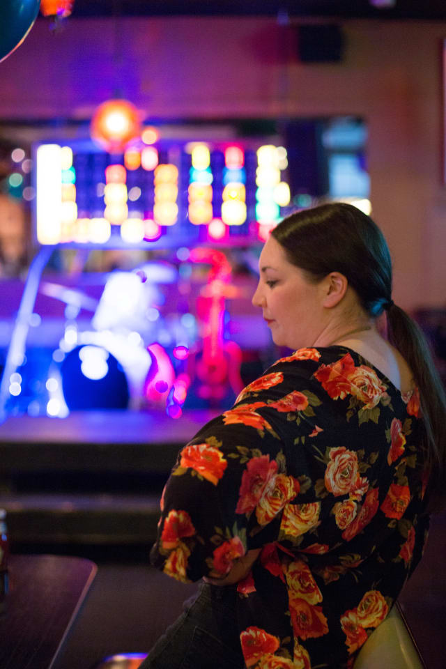 Woman in floral shirt seated at a bar, with colorful stage lights and musical instruments in the background, capturing the vibrant atmosphere of the Love Bomb 2017 event supporting My Voice Music.